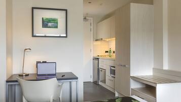 A modern hotel room with a bed, a desk with a laptop, a chair, a framed picture on the wall, and a kitchenette in the background.