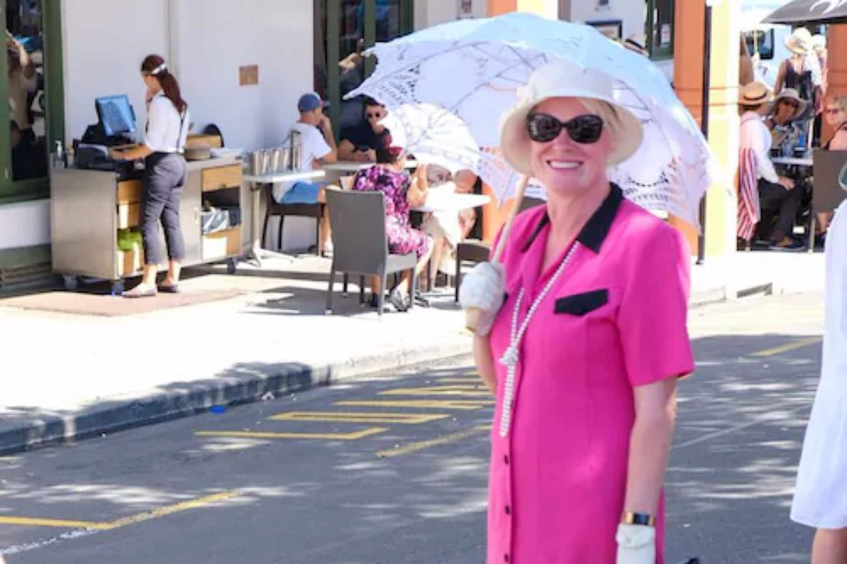 Una mujer con camisa rosa y sombrero blanco parada en una acera.