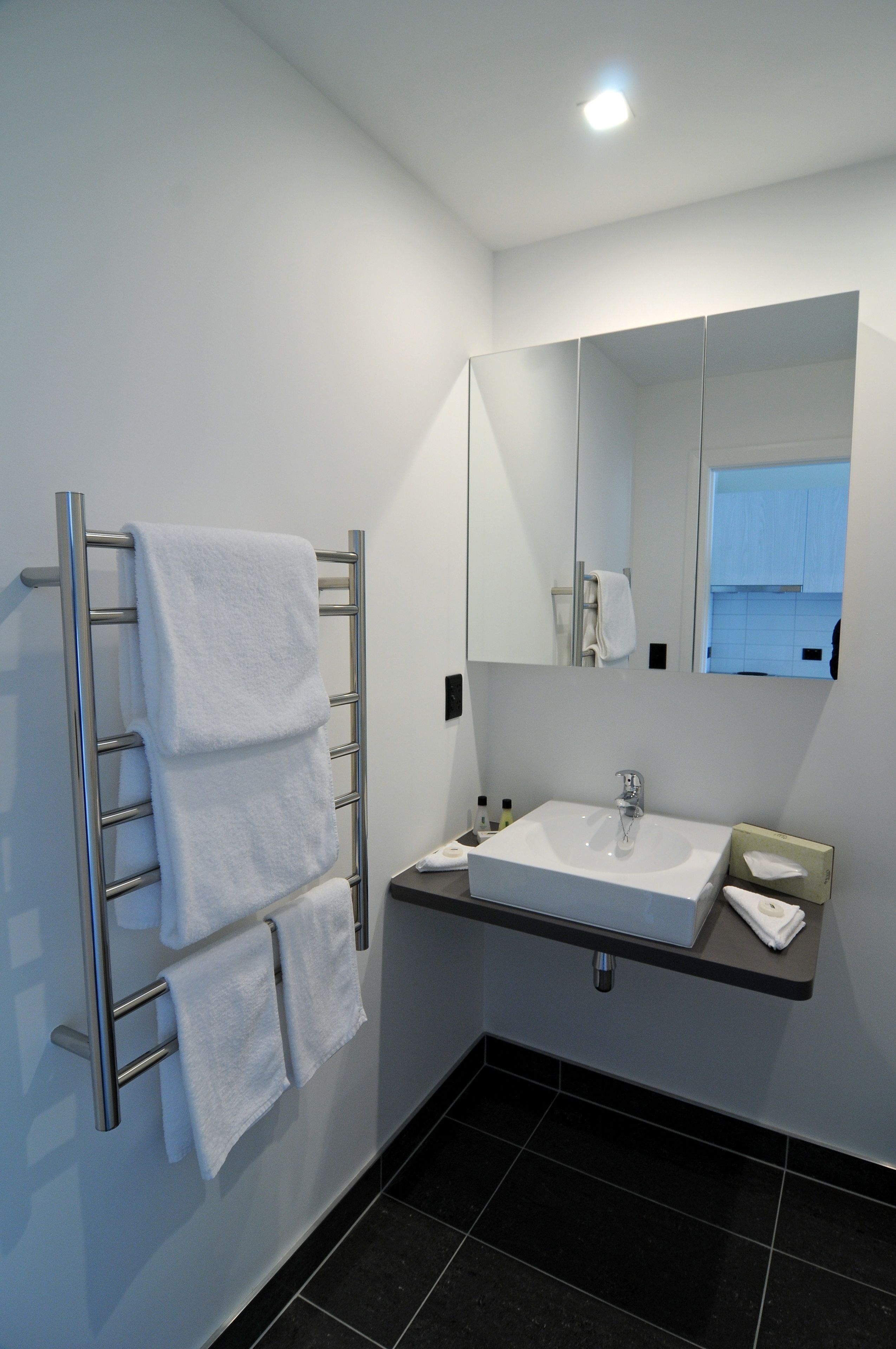 Un bany modern amb un lavabo integrat, una barra de tovalloles amb diverses tovalloles, un mirall i un sòl de rajola fosca.
