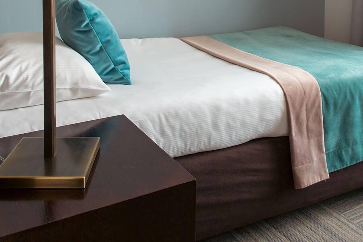 Ein ordentlich bezogenes Bett mit einem türkisfarbenen Kissen, einem weißen Kopfteil und einem braunen Bettrundenrock. Eine Stehlampe mit weißem Lampenschirm steht auf einem dunklen Nachttisch.