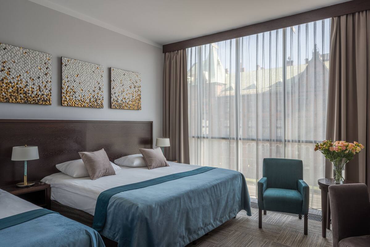 Ein Hotelzimmer mit einem großen Bett, zwei Nachttischen, einem Sessel, einem kleinen Tisch mit Blumenvase und Blick auf die Stadt durch durchsichtigen Vorhängen.