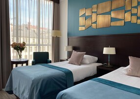 A hotel room with two beds, a view of buildings through large windows, and a decorative wall piece.