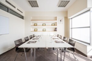 Une salle de conférence avec une longue table, des chaises, un tableau blanc, des étagères ornées de tableaux encadrés et une grande fenêtre.