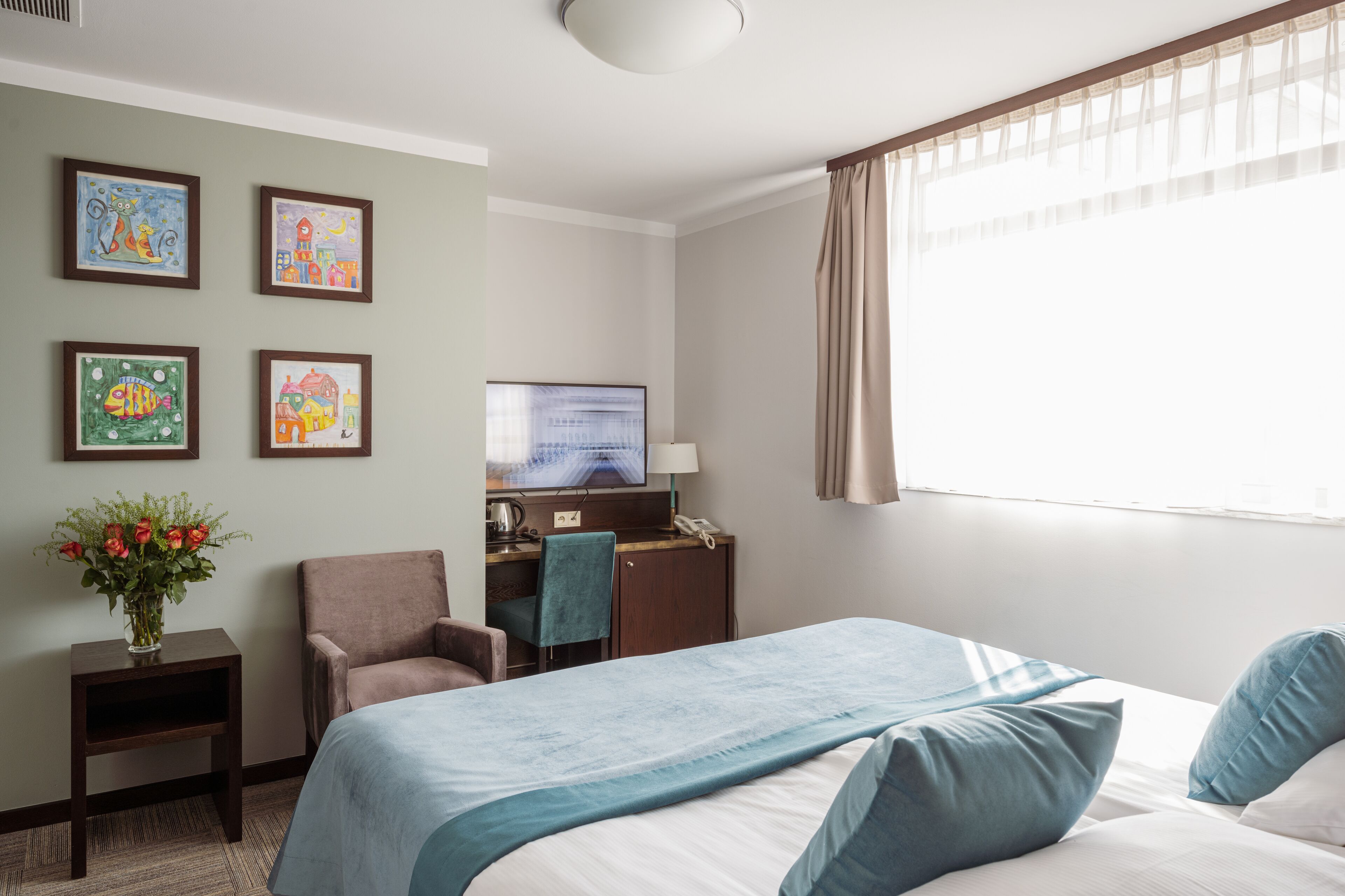 A hotel room with a bed, a desk, a chair, a TV, and artwork on the wall.