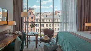 Une chambre d’hôtel avec une grande fenêtre, un bureau avec un téléphone, une chaise, une petite table avec un vase et un lit.
