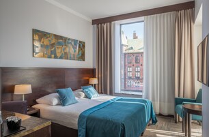 Habitación de hotel con una cama grande, dos mesitas de noche con lámparas, vista a un edificio por la ventana y un cuadro en la pared.