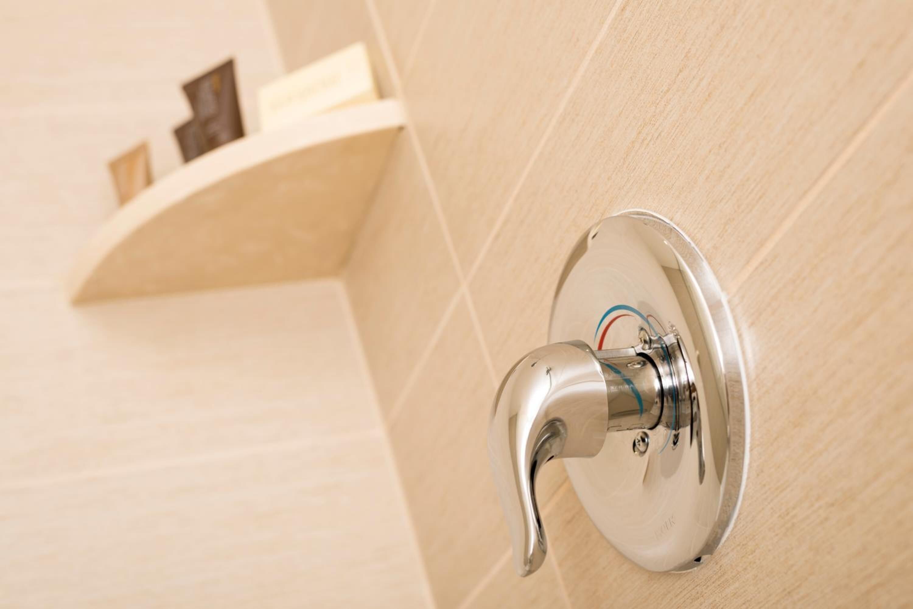 A bathroom with a wall-mounted shower faucet and a shelf with toiletries.