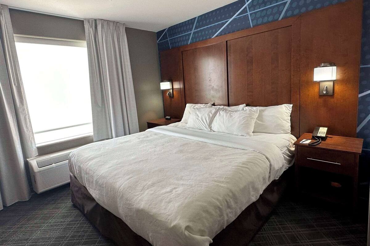 Une chambre d’hôtel avec un grand lit, deux tables de chevet avec des lampes, une fenêtre avec des rideaux et un mur orné d’un motif géométrique.
