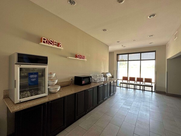 A modern kitchen with a refrigerator, microwave, and a coffee machine. There are two shelves with motivational words, and a large window with chairs outside. - Comfort Suites Jackson-Cape Girardeau (Jackson)