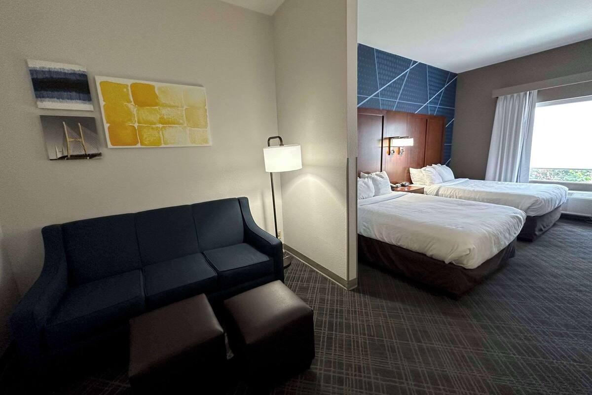 Une chambre d’hôtel avec un canapé de couleur sombre, un lampadaire, deux lits et une grande fenêtre avec des rideaux.