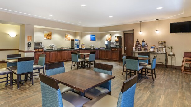 A hotel dining area with multiple tables and chairs, a kitchen area with a 'WASTE' sign, and a television mounted on the wall.