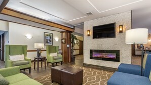 A hotel lobby with green armchairs, a brown ottoman, a fireplace, and a television mounted on the wall.