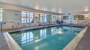 An indoor swimming pool with clear glass walls, white lounge chairs, and a tiled deck.