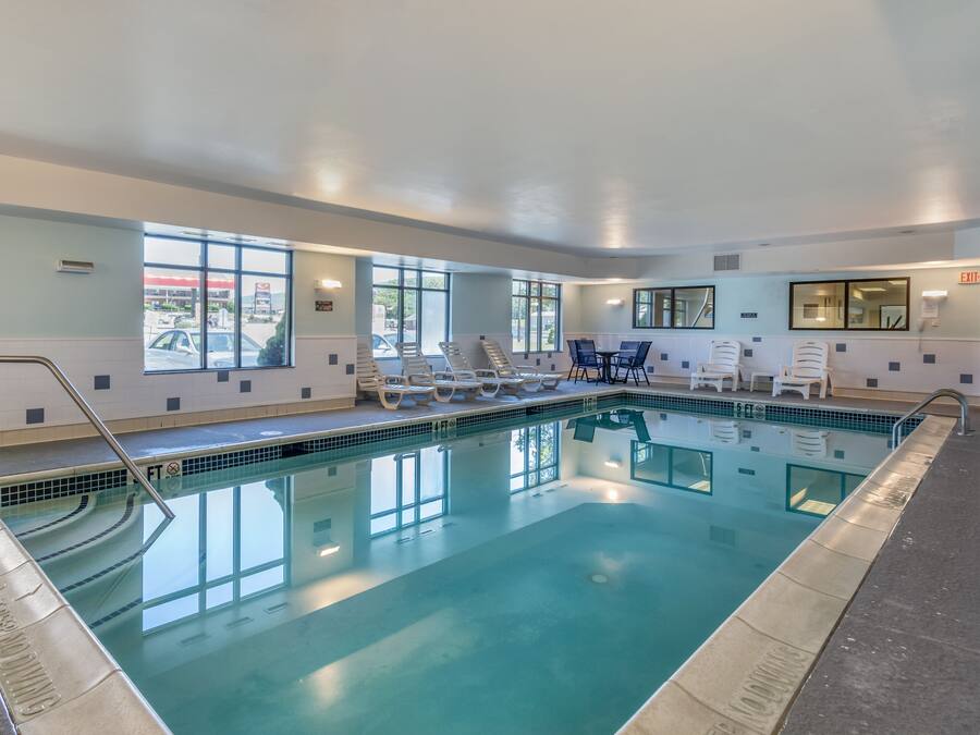 An indoor swimming pool with clear glass walls, white lounge chairs, and a tiled deck.