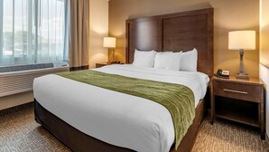 A hotel room with a large bed, a wooden headboard, two bedside tables with lamps, a window with sheer curtains, and a carpeted floor.