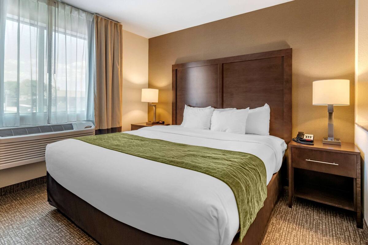Una habitación de hotel con una cama grande, un cabecero de madera, dos mesitas de noche con lámparas, una ventana con cortinas de encaje y suelo alfombrado.