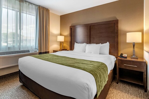 A hotel room with a large bed, a wooden headboard, two bedside tables with lamps, a window with sheer curtains, and a carpeted floor.