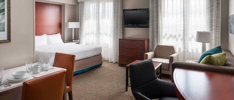 A hotel room with a bed, a desk with chairs, a television, and a dining area.