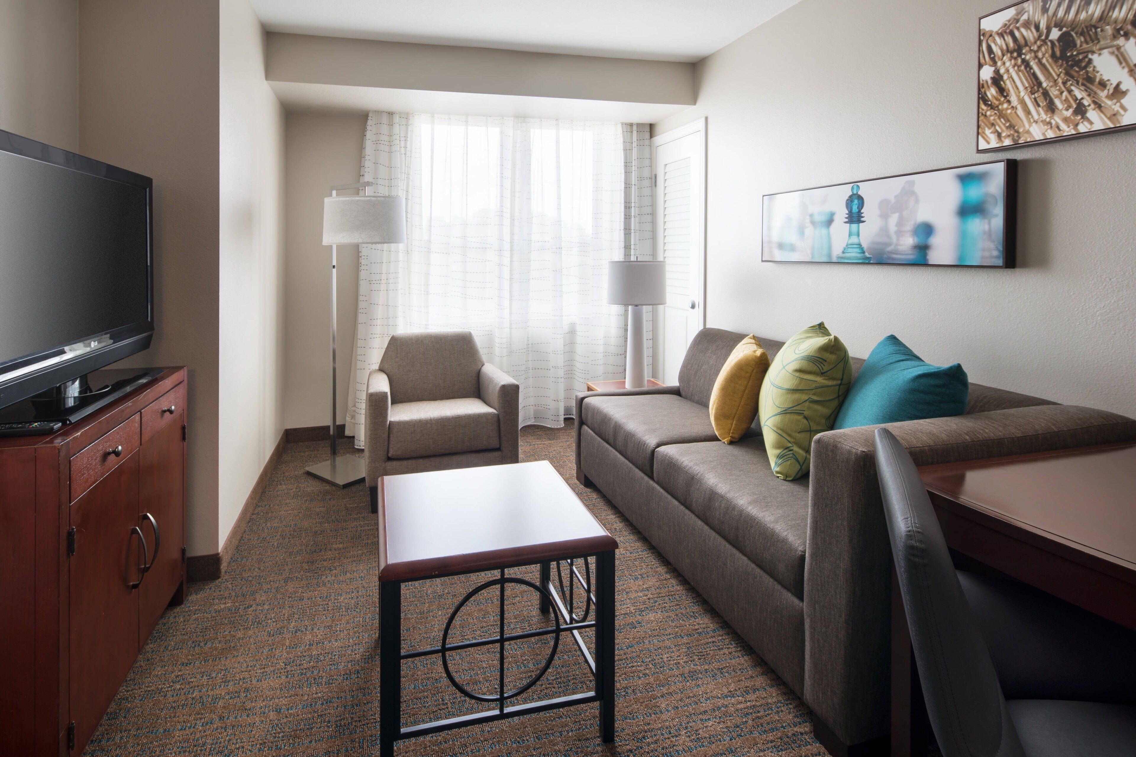 Habitación de hotel con televisión de pantalla plana, sofá con cojines, una mesita, lámpara y ventana con cortinas traslúcidas.