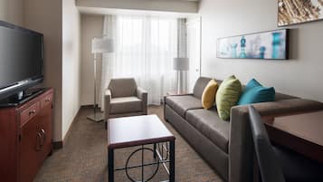 Habitación de hotel con televisión de pantalla plana, sofá con cojines, una mesita, lámpara y ventana con cortinas traslúcidas.