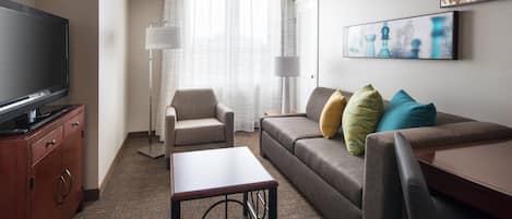 Habitación de hotel con televisión de pantalla plana, sofá con cojines, una mesita, lámpara y ventana con cortinas traslúcidas.