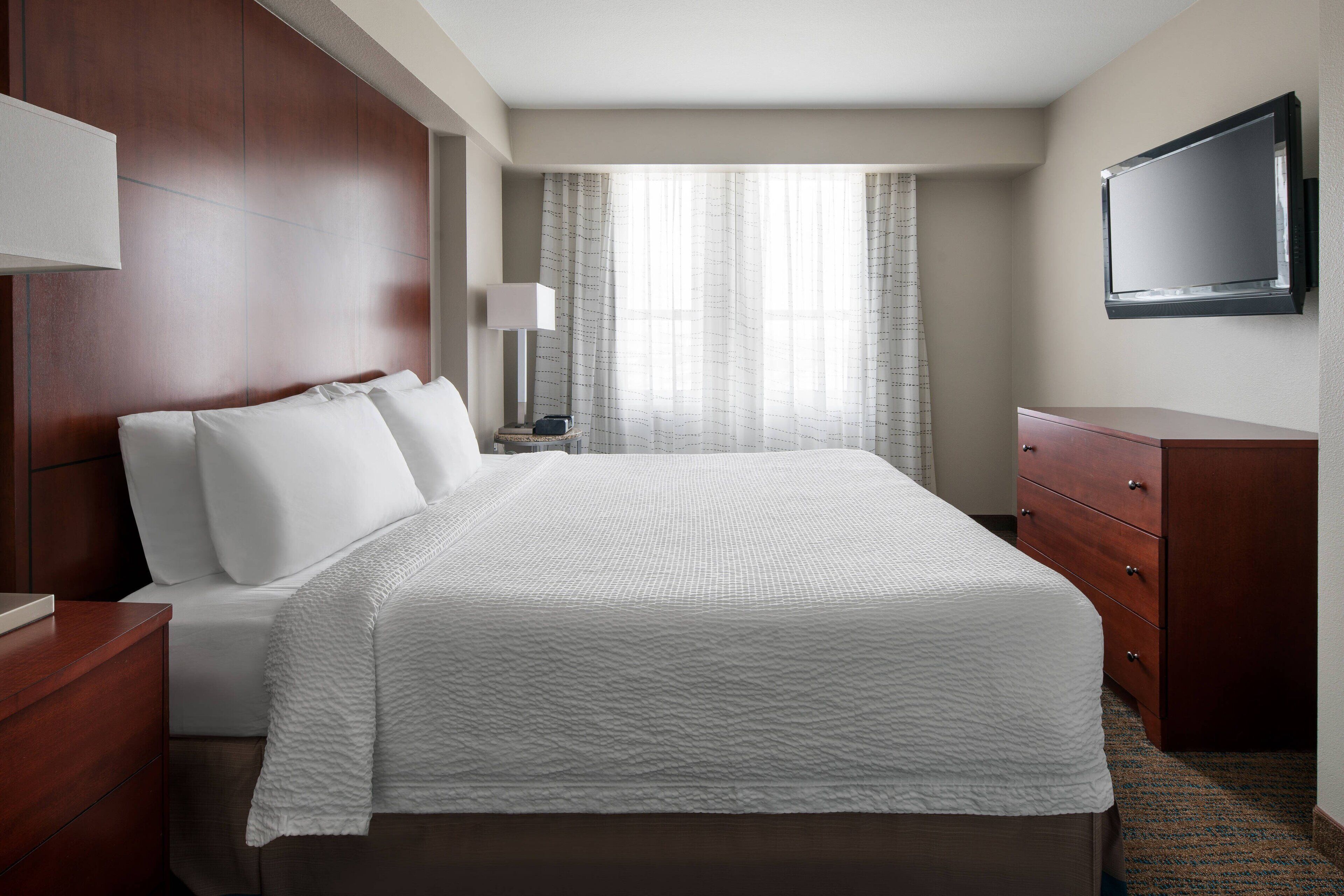 Une chambre d’hôtel avec un grand lit, une commode en bois, une table de chevet, une lampe et un téléviseur à écran plat fixé au mur.