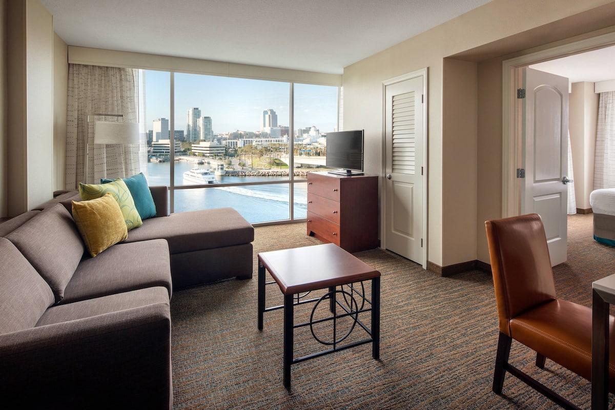 A hotel room with a large window, a sofa, a television, a small table, and a chair.