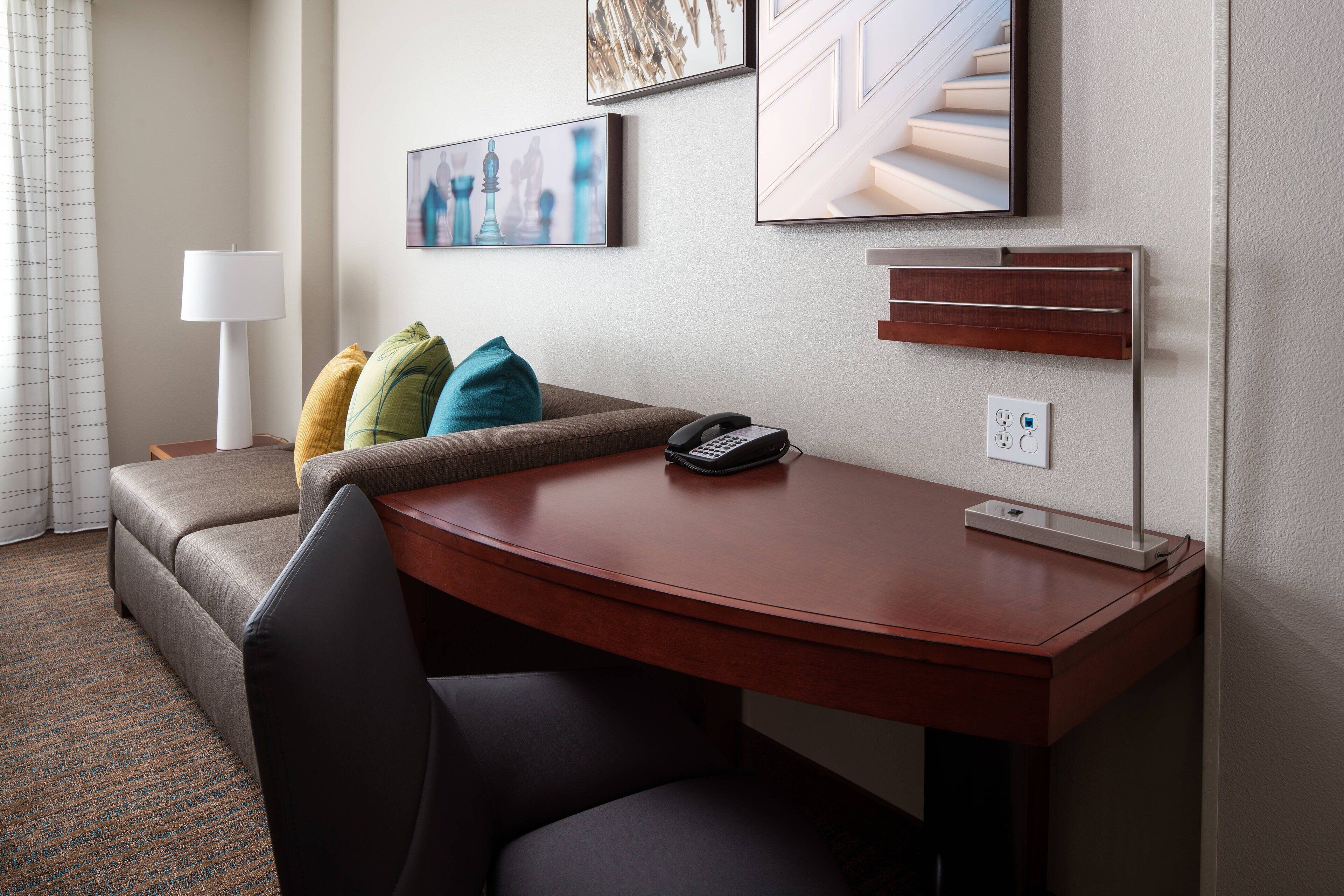 A hotel room with a sofa, a desk, a lamp, a phone, and artwork on the wall.