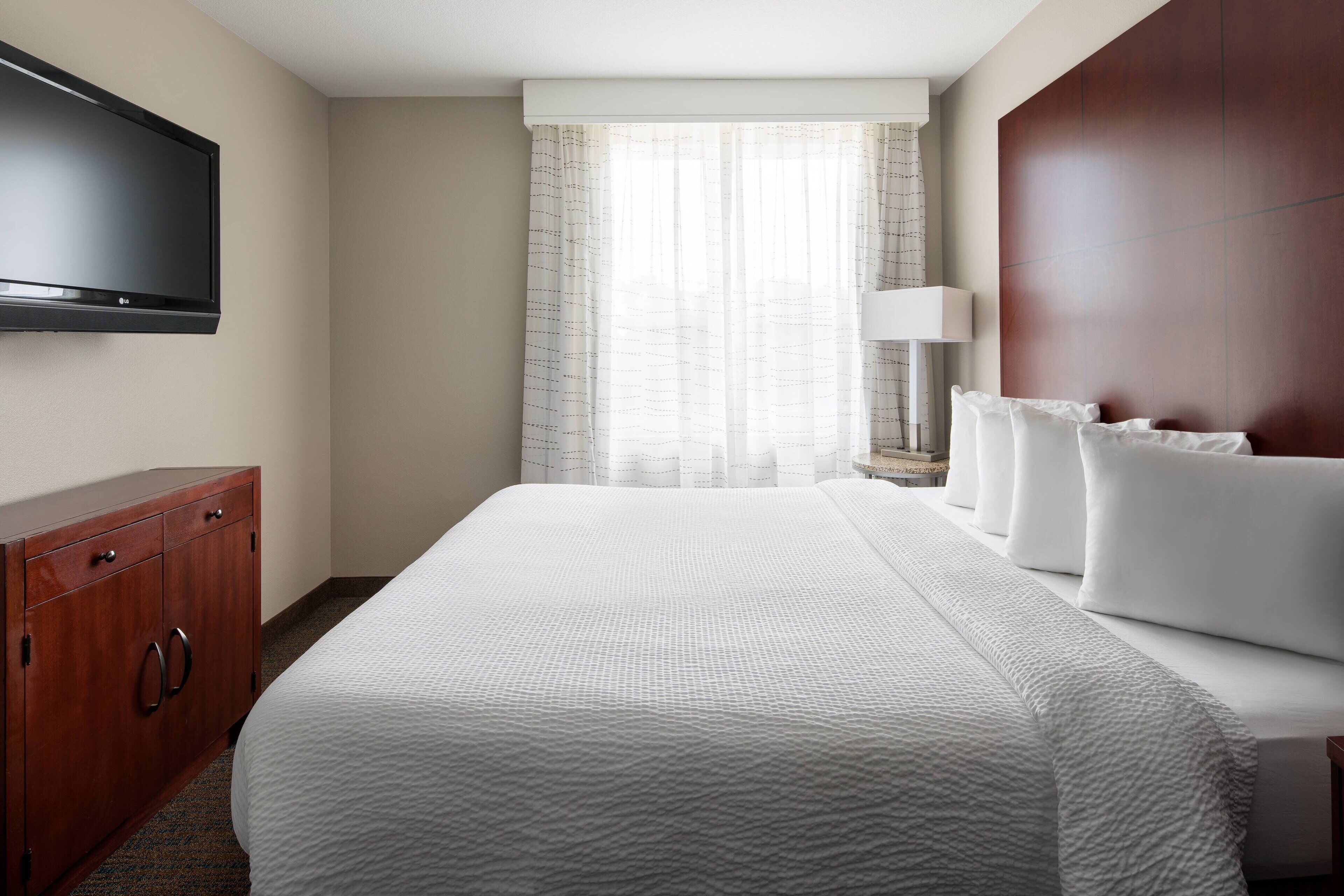A hotel room with a large bed, a flat-screen TV mounted on the wall, a wooden dresser, and a window with sheer curtains.