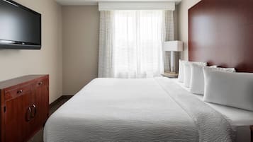 Habitación de hotel con una cama grande, un televisor de pantalla plana montado en la pared, un aparador de madera y una ventana con cortinas traslúcidas.