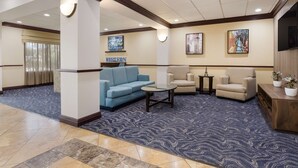 A lobby with a blue patterned carpet, a blue sofa, beige armchairs, a small round table, and a wooden console table with potted plants. - Best Western Coffeyville Central Business District Inn and Suites (Coffeyville)