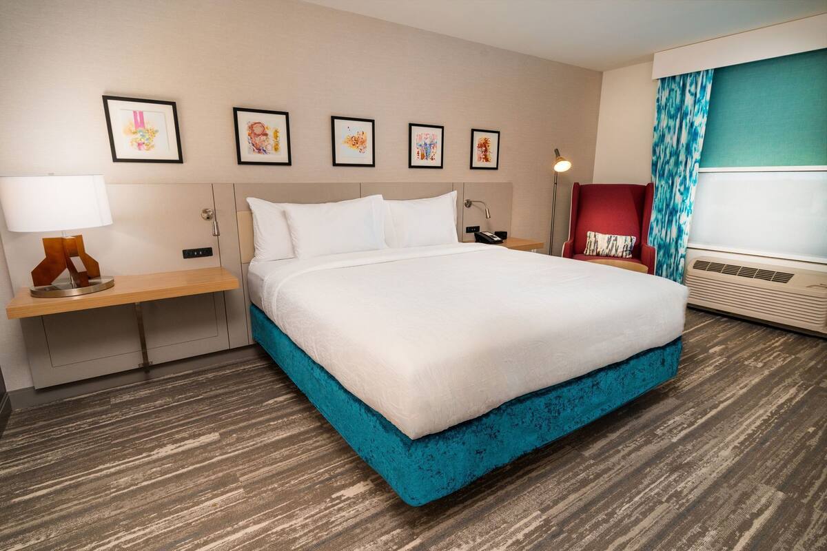 A hotel room with a bed, bedside tables, a desk, a lamp, a chair, a heater, and artwork on the wall.
