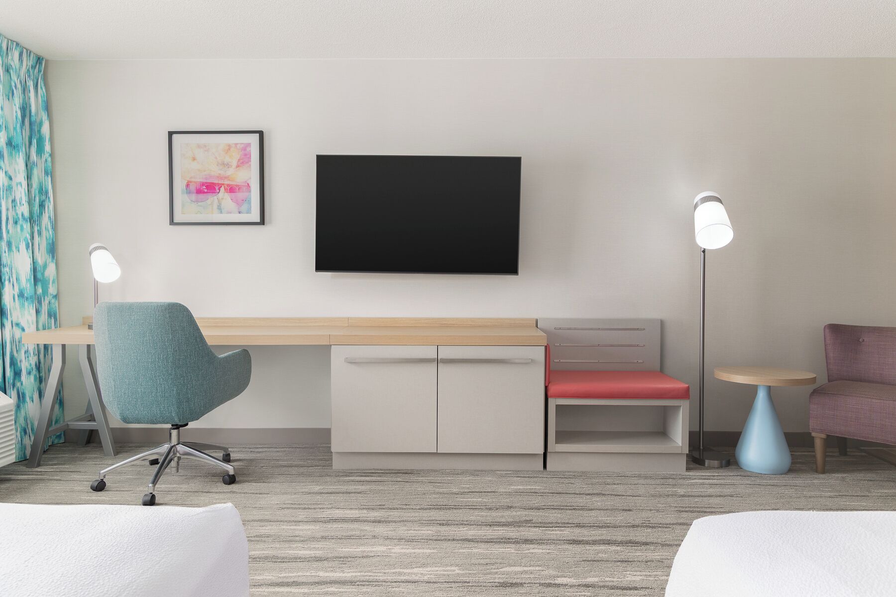 A modern hotel room with a desk, a flat-screen TV, a red chair, a blue chair, and two floor lamps.