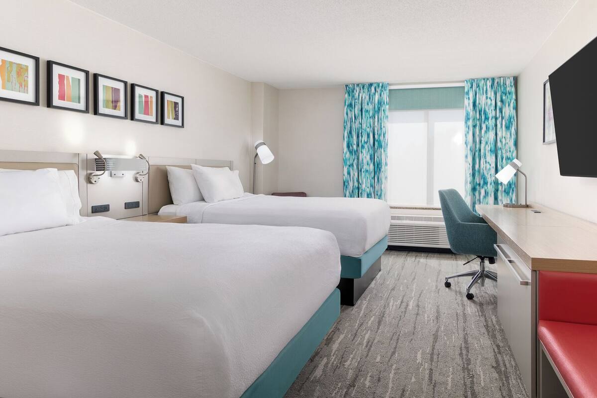 A hotel room with two beds, a desk, a chair, a TV, and a window with curtains.