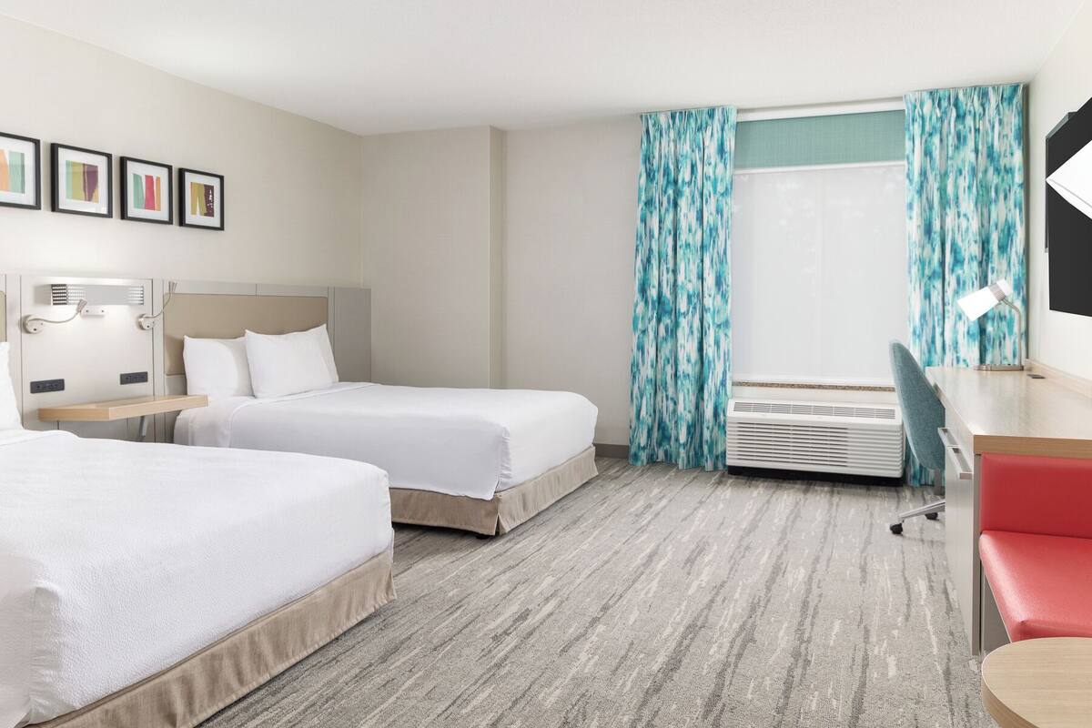 A hotel room with two beds, a desk, a chair, a desk lamp, and a window with curtains.