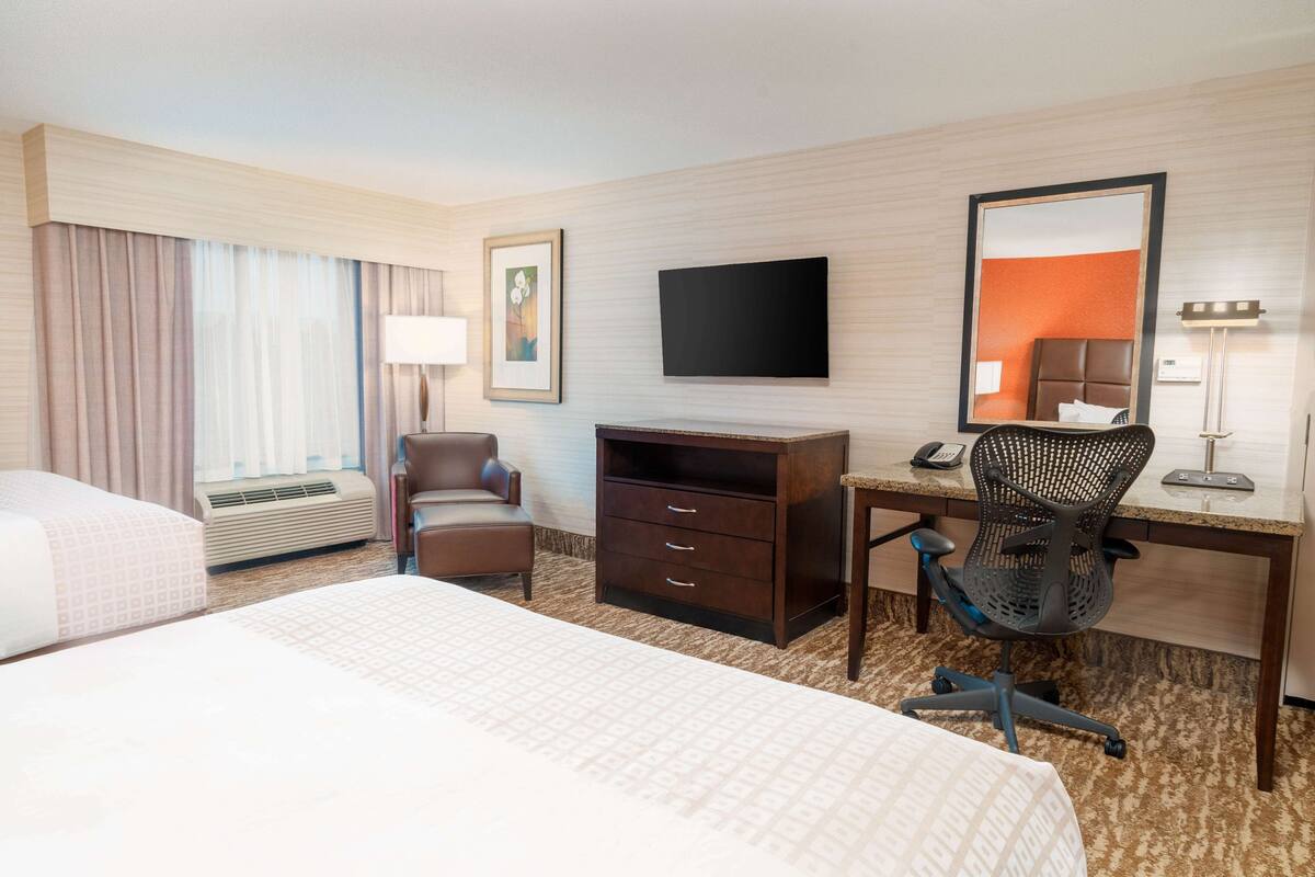 A hotel room with two beds, a desk with a chair, a flat-screen TV, a dresser, and a large mirror.