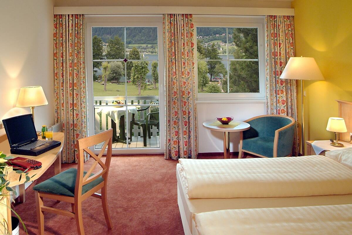 Habitación de hotel con una cama, un escritorio con una computadora portátil, una silla, una mesita con fruta y vistas a la orilla de un lago.