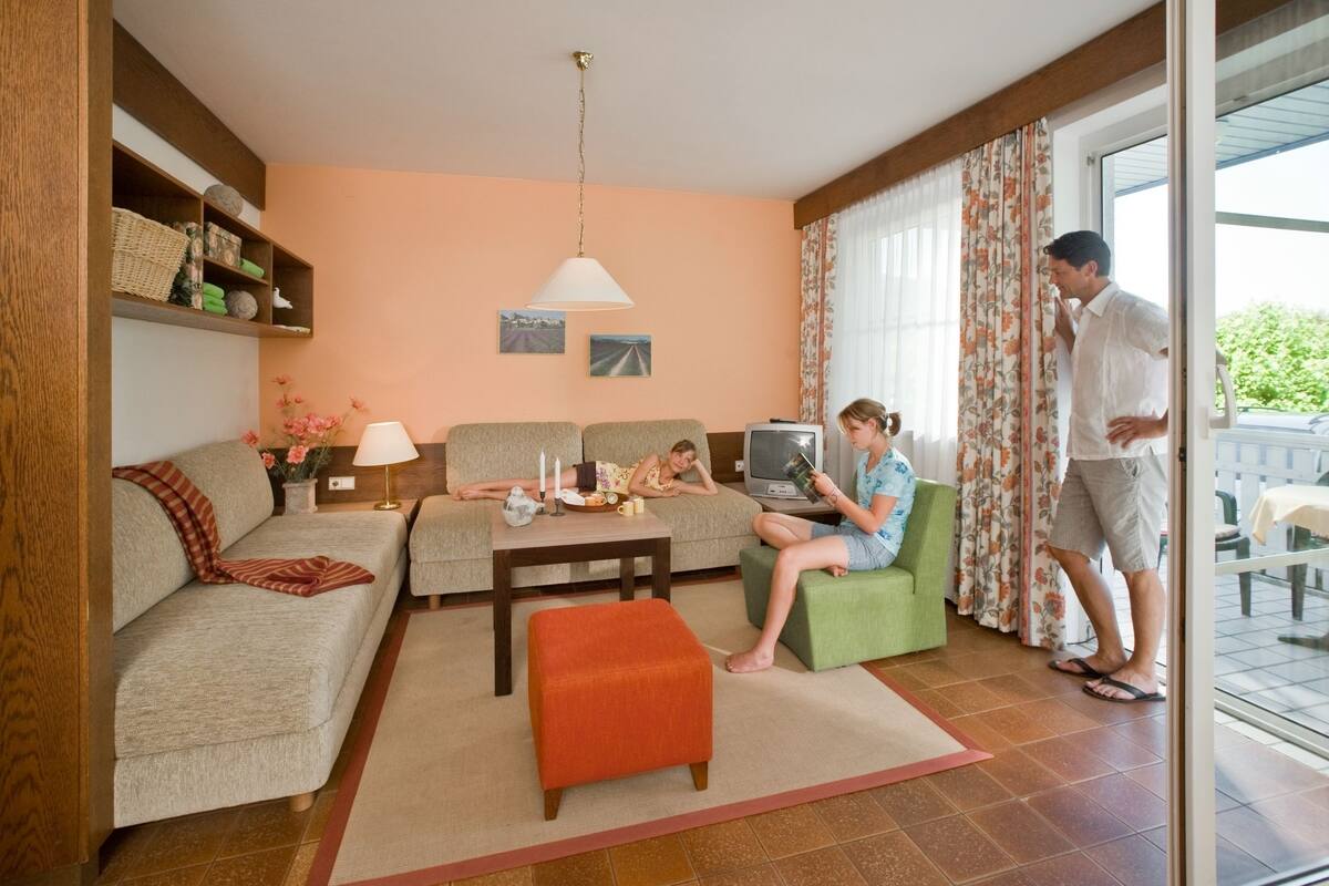 Una sala de estar acogedora con un sofá, un reposapiés y un televisor. Dos personas sentadas y una de pie junto a la ventana.