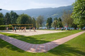 A volleyball court with a net, surrounded by a grassy area and trees, with mountains in the background.