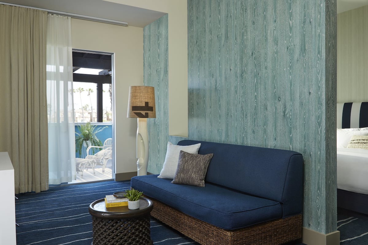 A hotel room with a blue sofa, a small round table with a plant, and a view of palm trees through a sliding door.