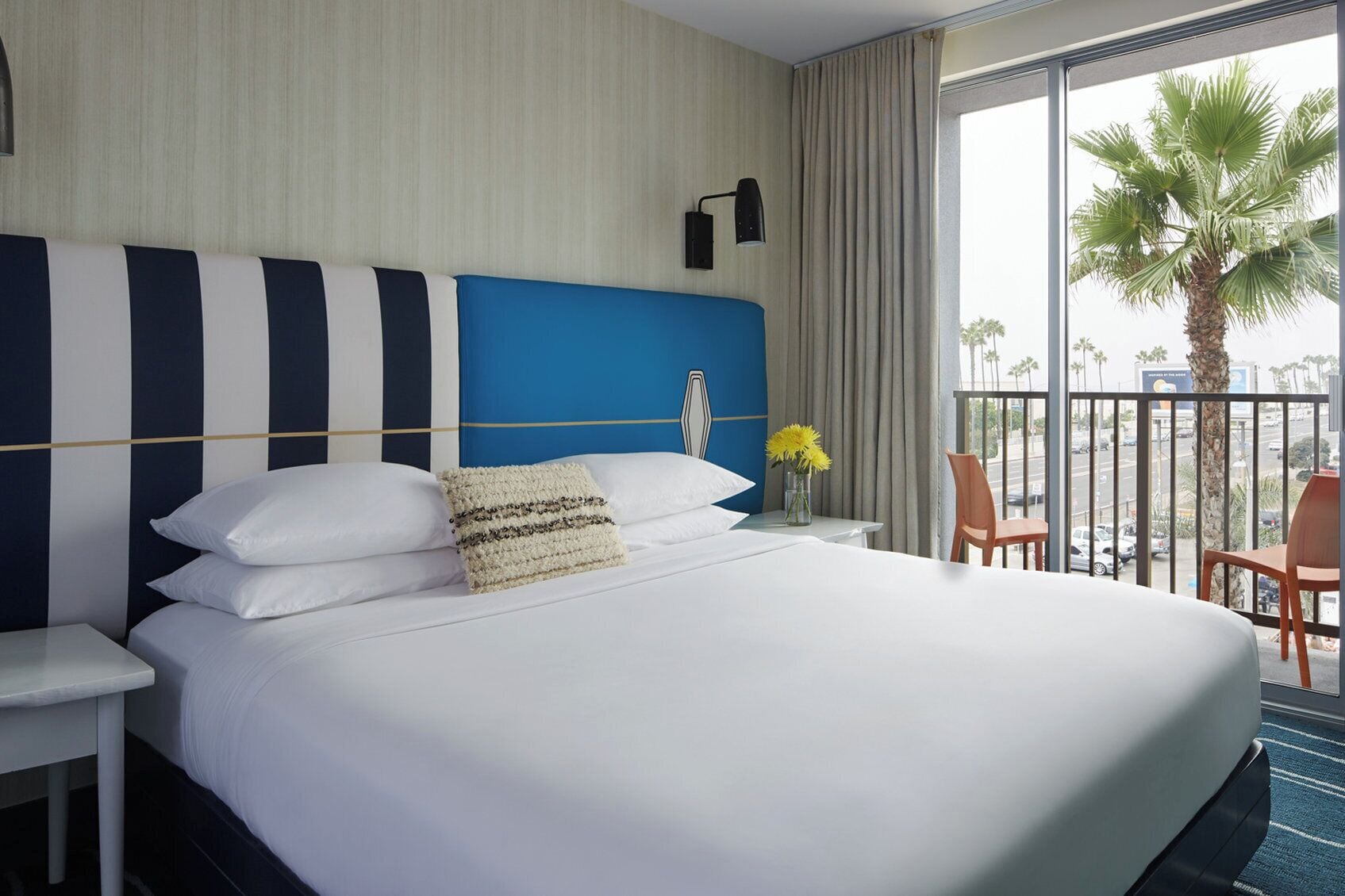 Une chambre d’hôtel avec un grand lit, une table de chevet, une lampe, une chaise et une vue sur les palmiers depuis un balcon.