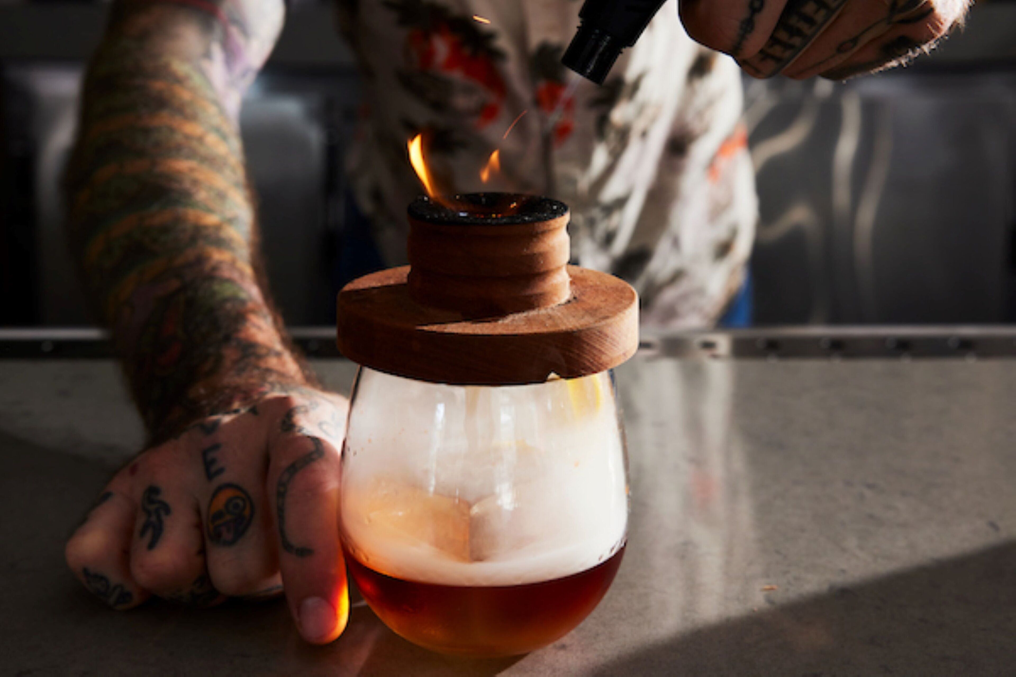 A person with tattoos lighting a candle in a glass container.