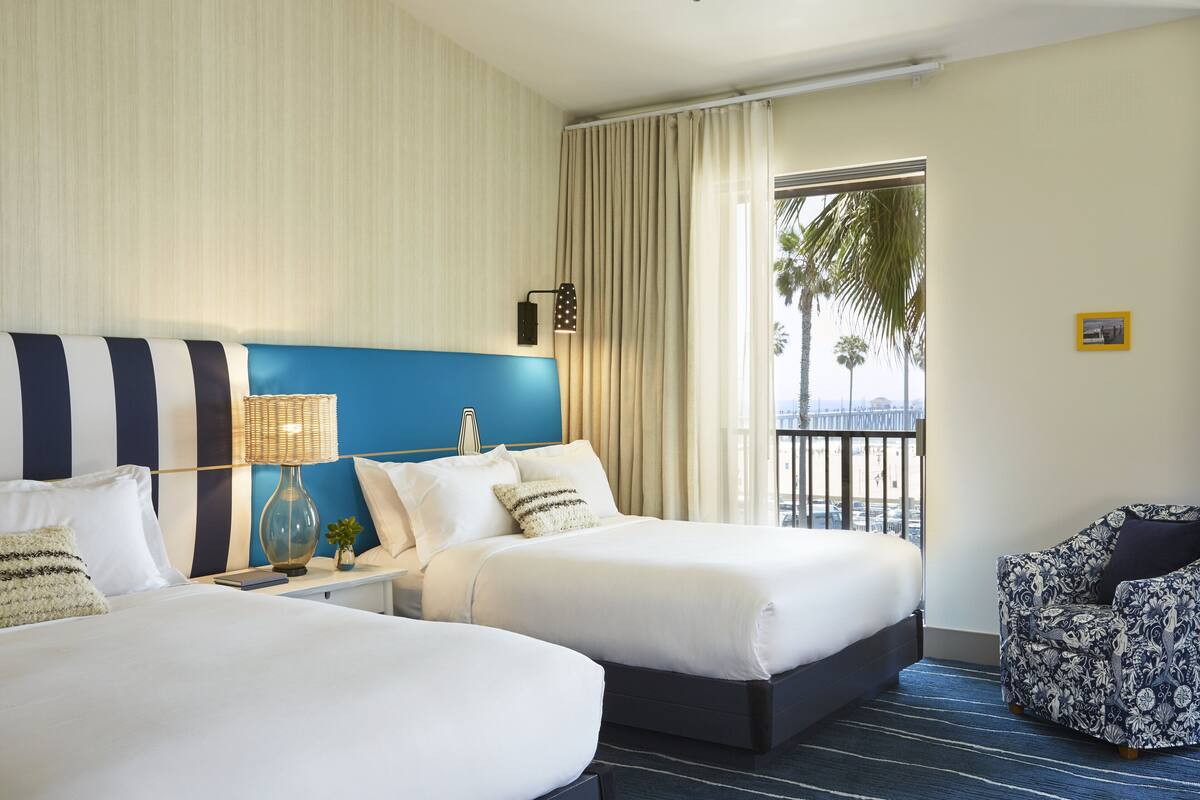 A hotel room with two beds, a nightstand, a lamp, a chair, and a view of palm trees through a window.