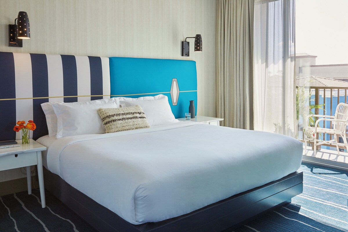 A hotel room with a large bed, a nightstand, a lamp, and a view of the outside.