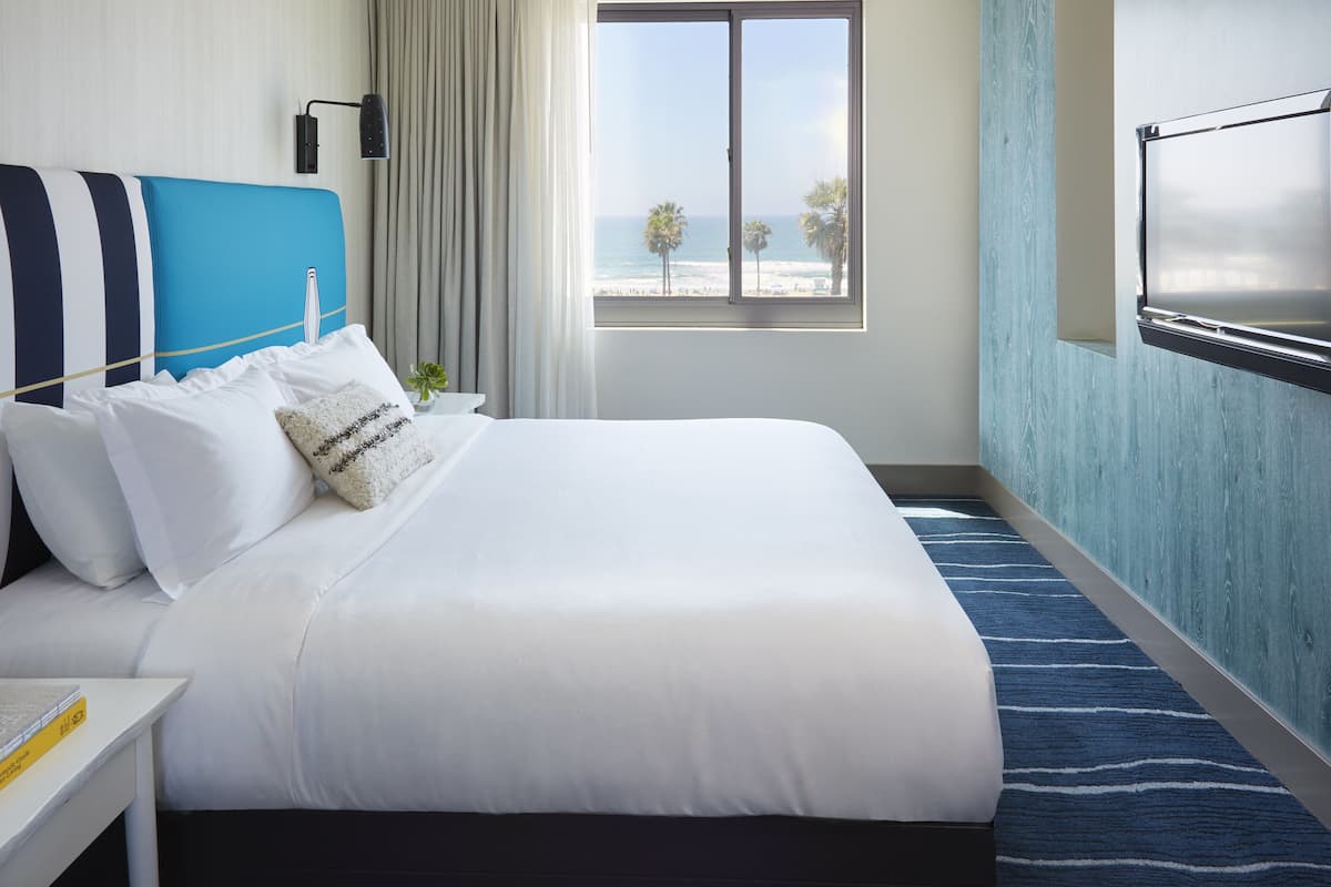 A hotel room with a large bed, a TV, a window with a view of the beach, and a nightstand with a lamp and a plant.