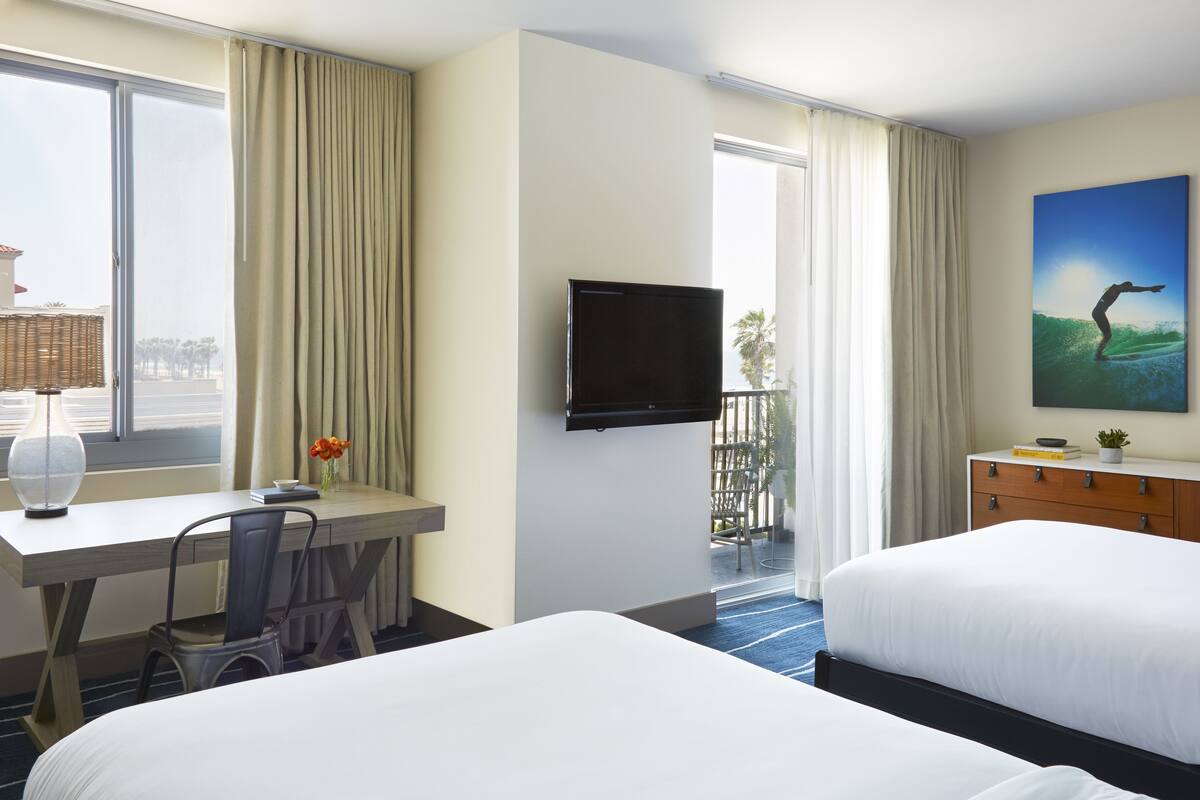 A hotel room with two beds, a desk with a chair, a television mounted on the wall, and a window with curtains.