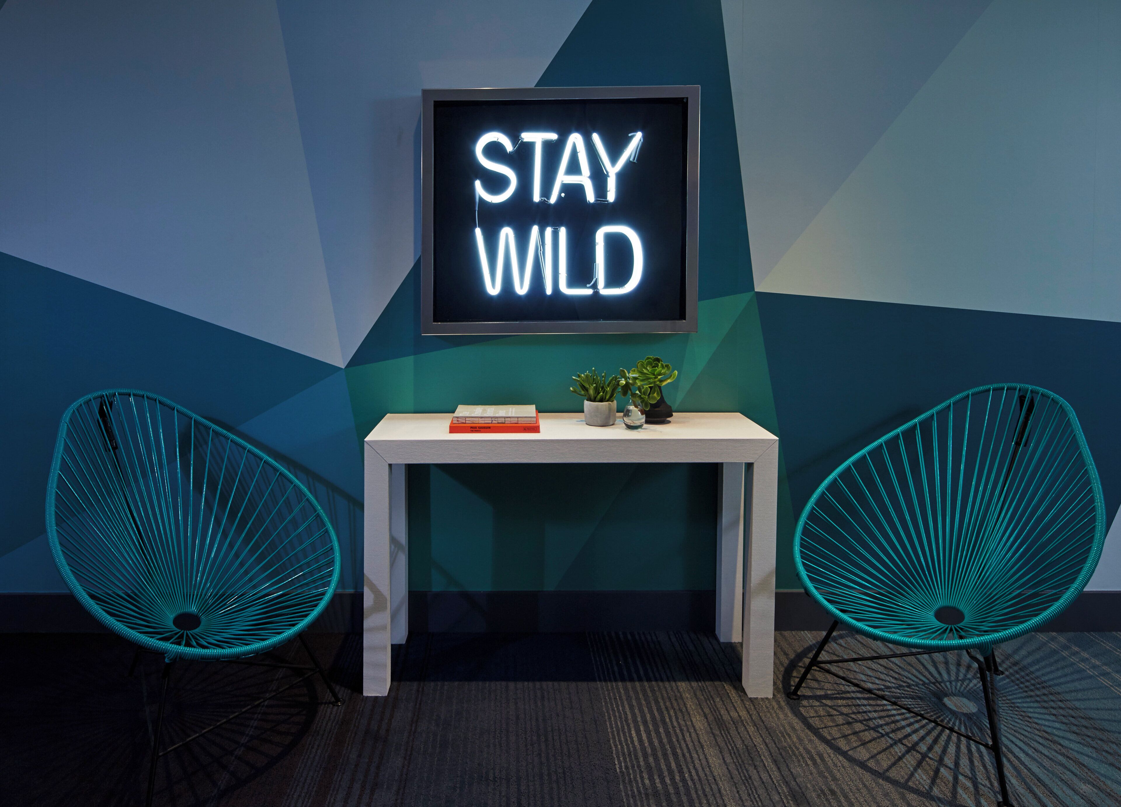 A room with a neon sign that reads 'STAY WILD', a small table with a book and two potted plants, and two teal chairs.