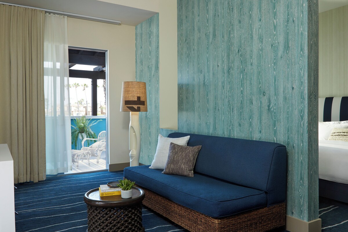 A hotel room with a blue sofa, a small table with a plant, a lamp, and a view of the outdoors.