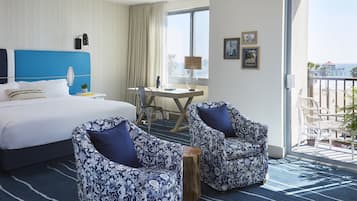 Ett hotellrum med en stor säng, två mönstrade fåtöljer, ett skrivbord med en lampa och utsikt över stranden från balkongen.