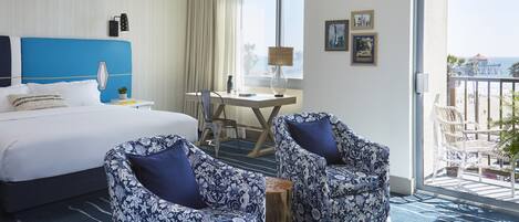 A hotel room with a large bed, two patterned armchairs, a desk with a lamp, and a view of the beach through a balcony.